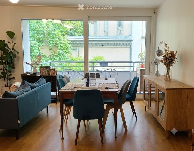 Open-plan living and dining area with wooden flooring, large windows, and a balcony. Features a blue sofa, dining table with chairs, a wooden cabinet, and decorative items.