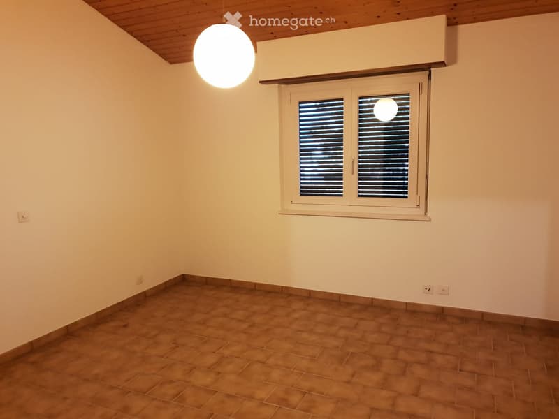 Empty room with beige tiled floor, white walls, and a single window with shutters. A round pendant light hangs from the ceiling.