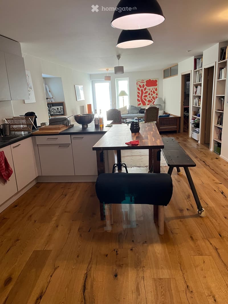 Open-plan living and dining area with wooden flooring, a large central dining table, a sofa in the background, and a kitchen area with cabinets and a sink.