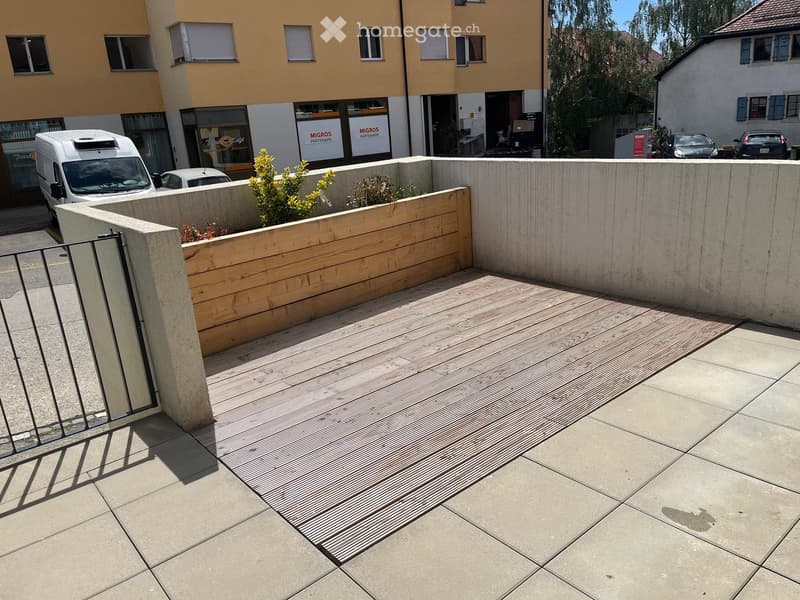 A modern outdoor patio with a wooden deck, concrete walls, and small plants. The area is enclosed by a metal gate and features a tiled floor.