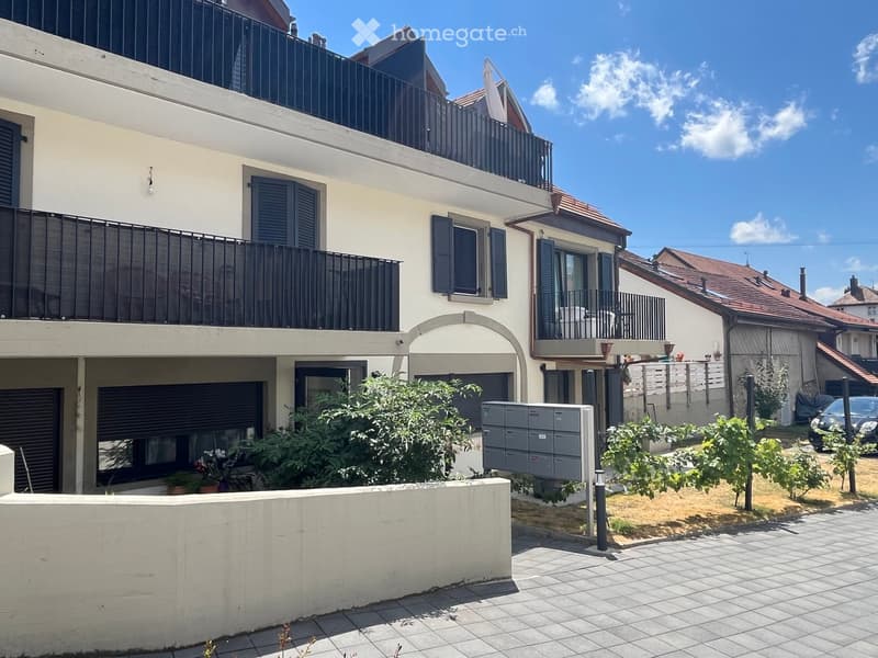 Two-story residential building with balconies, shutters, and a small garden area with potted plants and a mailbox.