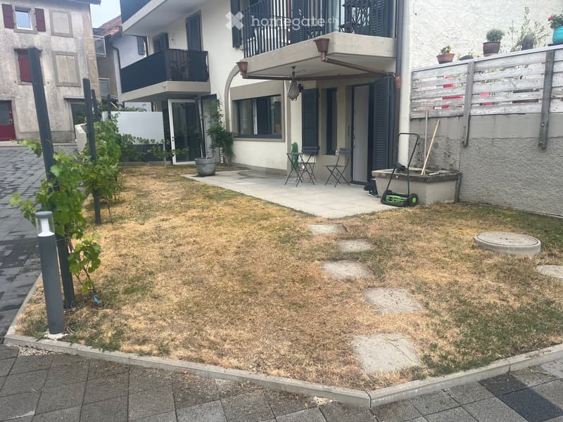 Garden area with dry grass, small tree, patio with chairs, and a building with balconies in the background.