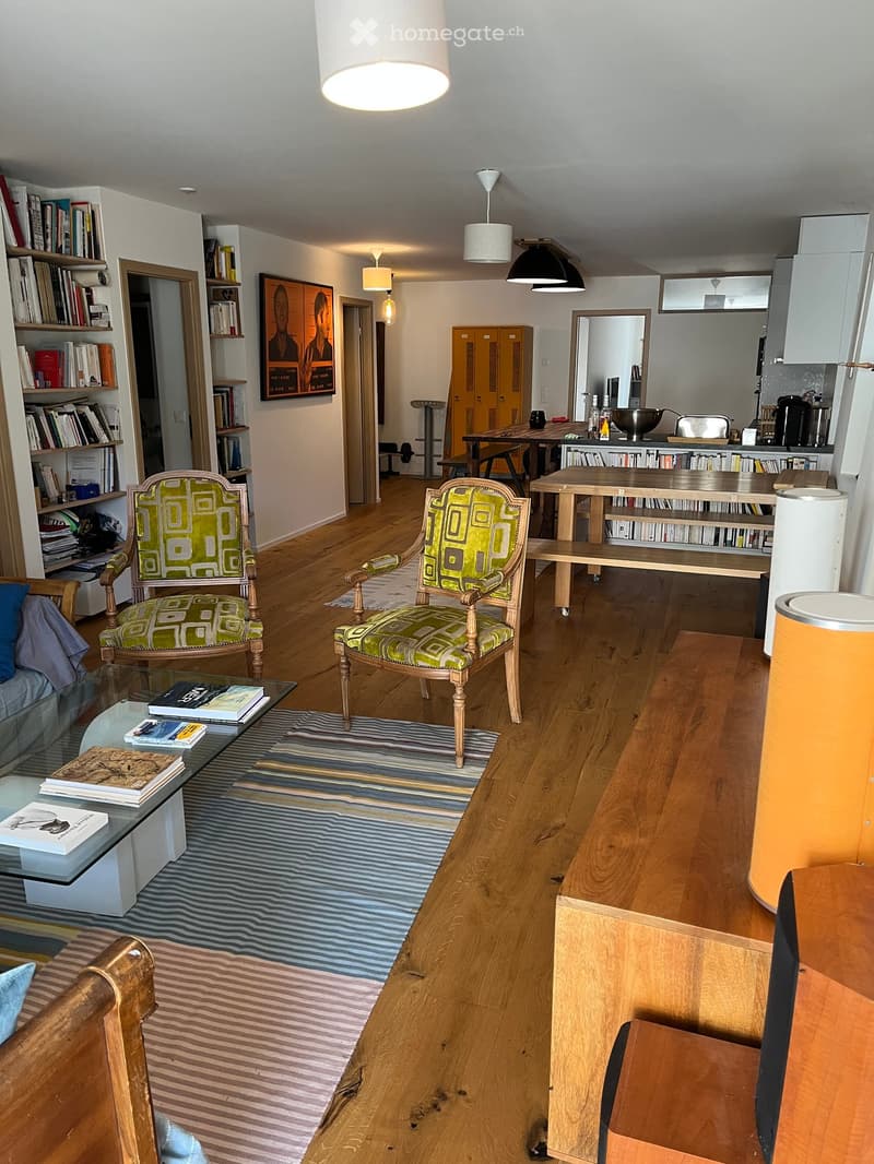 Open-plan living and dining area with wooden floors, modern furniture, and bookshelves. Features a glass coffee table, two patterned armchairs, a sofa, and a dining table with chairs.