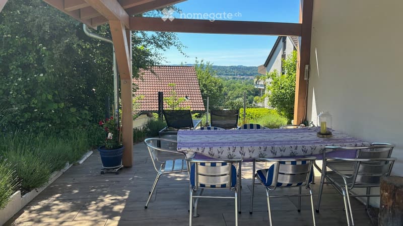 Wooden terrace with a table and chairs, overlooking a scenic landscape with trees and hills.