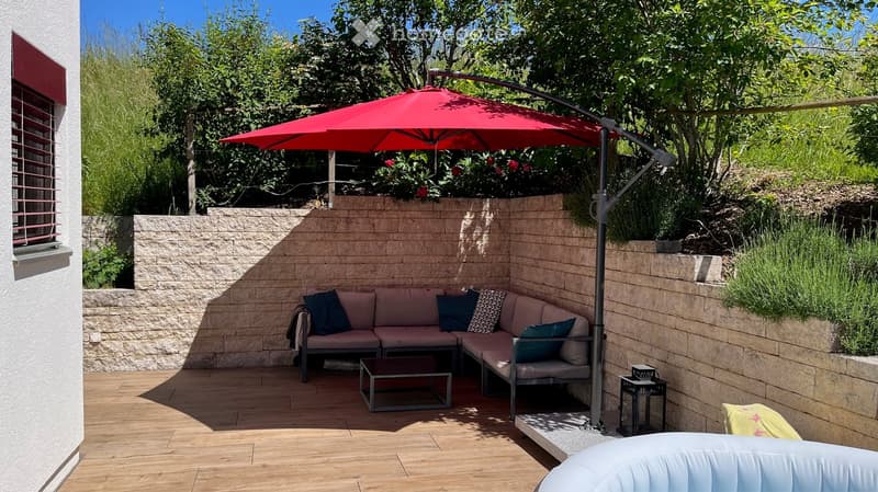 A modern outdoor seating area with a wooden deck, a grey sectional sofa, throw pillows, a coffee table, and a large red umbrella providing shade. The space is enclosed by a stone wall and features lush greenery and plants.