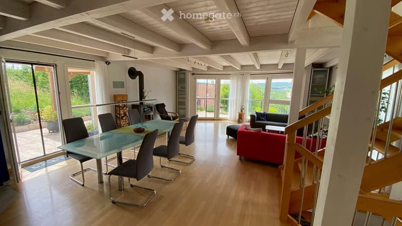 Bright, open-plan living and dining area with wooden floors, large windows, a red sofa, a glass dining table with chairs, and a fireplace.