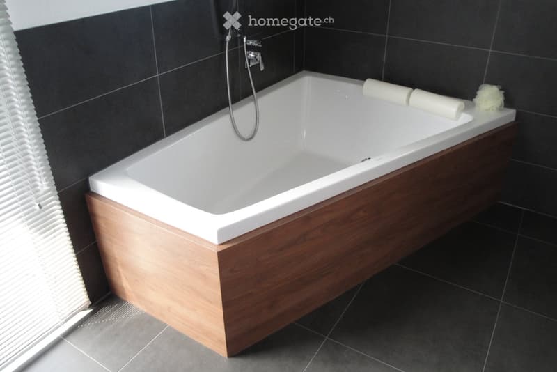 Modern bathroom with a white bathtub, wooden tray, and towels placed on the edge. Dark tiled walls and floor, with a window with blinds on the left side.