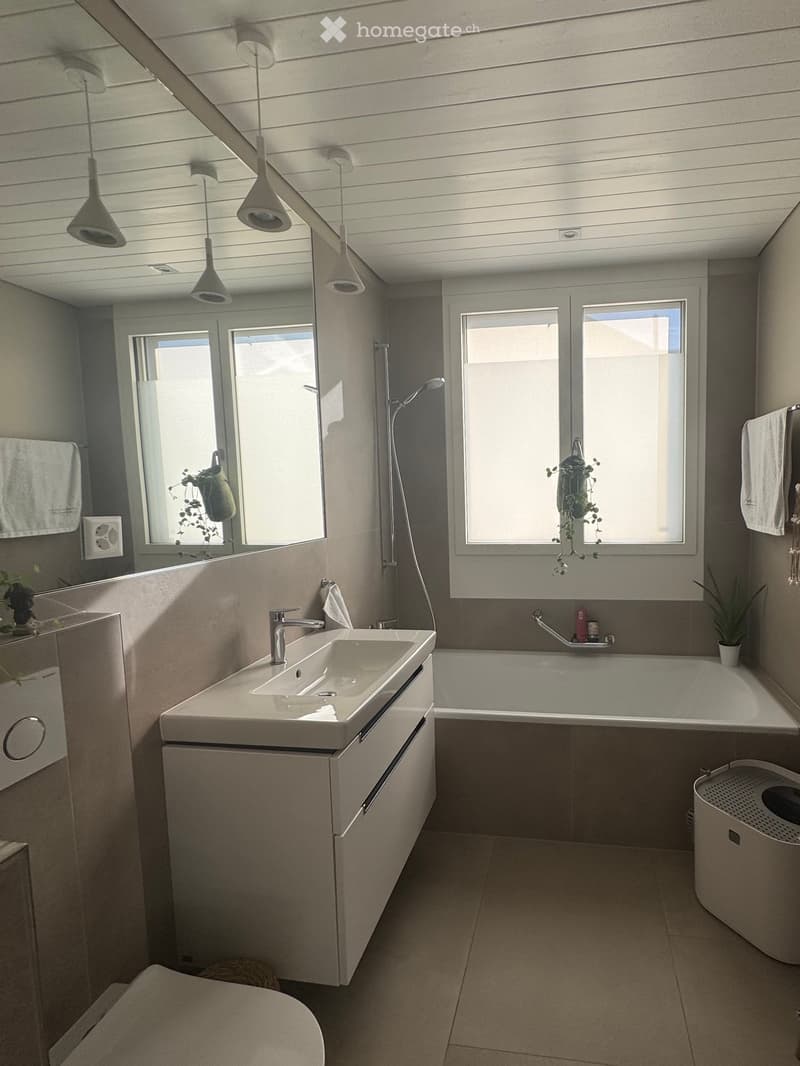 Modern bathroom with a white sink, bathtub, toilet, and large windows. Features a sleek faucet, modern light fixtures, and a mirror above the sink.