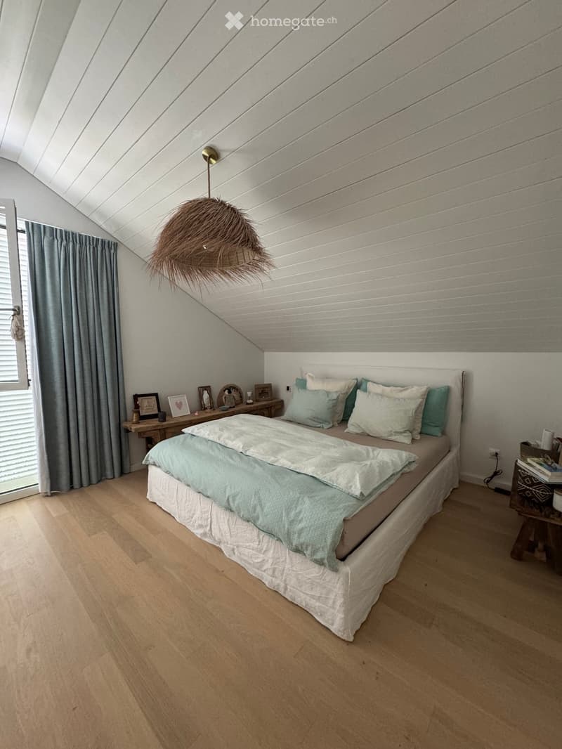 Cozy bedroom with a slanted wooden ceiling, a large bed with light blue bedding, a wooden nightstand, and a hanging woven lamp.
