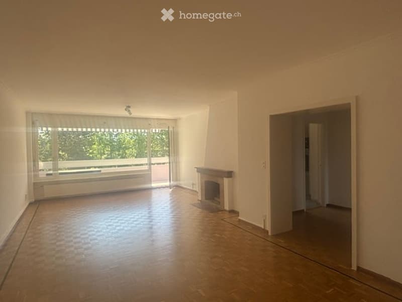 Empty room with wooden flooring, white walls, large window with blinds, fireplace, and access to a balcony.