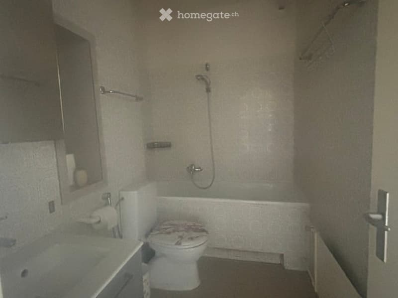 Bathroom with a toilet, bathtub, sink, mirror, and towel rack. The walls are covered in a light, patterned wallpaper, and the floor is dark.