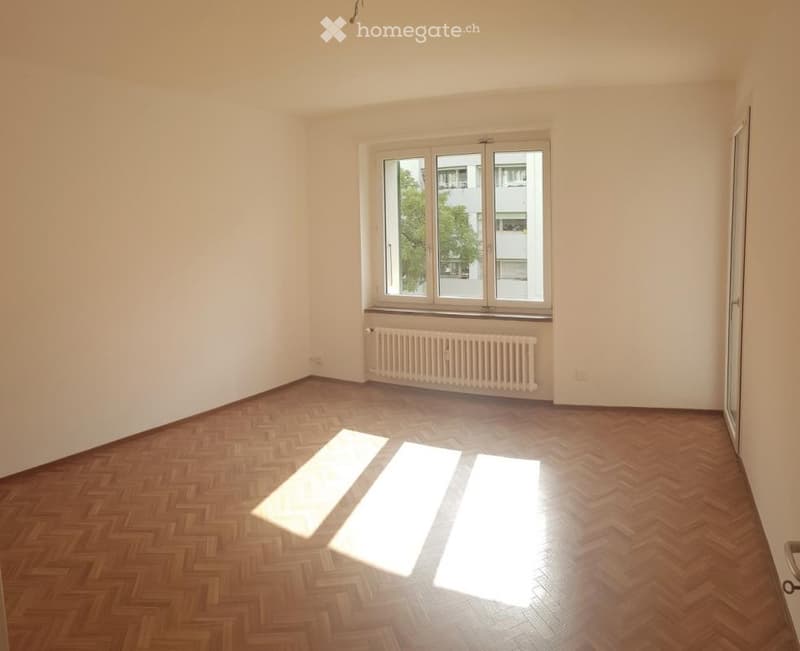 Empty room with wooden herringbone flooring, white walls, and a large window with a radiator below it.