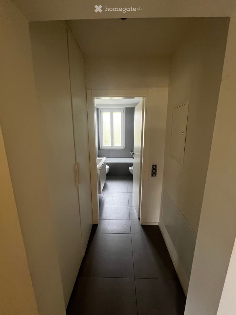 A narrow hallway with light-colored walls and dark tiled flooring, leading to a small bathroom with a window, sink, and toilet.