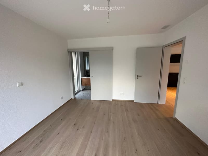 Empty room with light wooden flooring and white walls, featuring two open doors leading to other rooms.