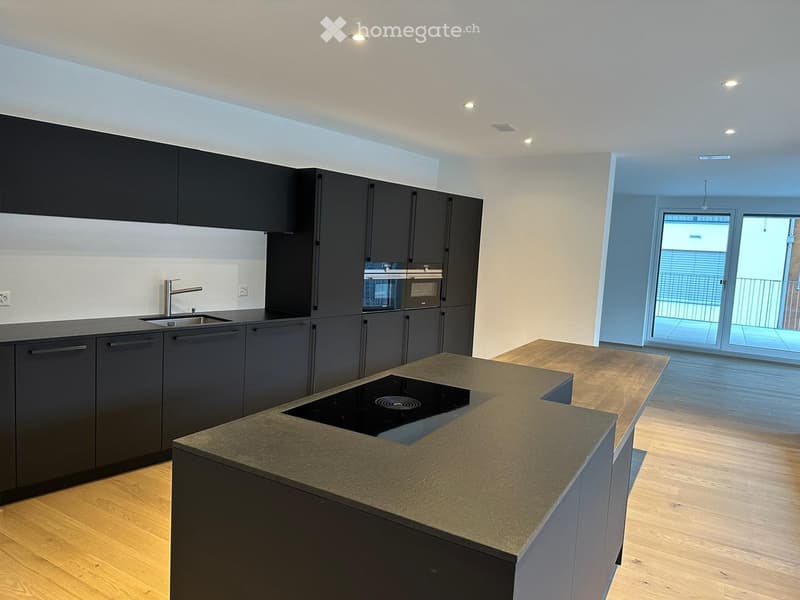 Modern kitchen with dark grey cabinets, a central island with an induction cooktop, and built-in appliances including an oven and microwave. The space features a sleek, minimalist design with a wooden floor and large windows leading to a balcony.
