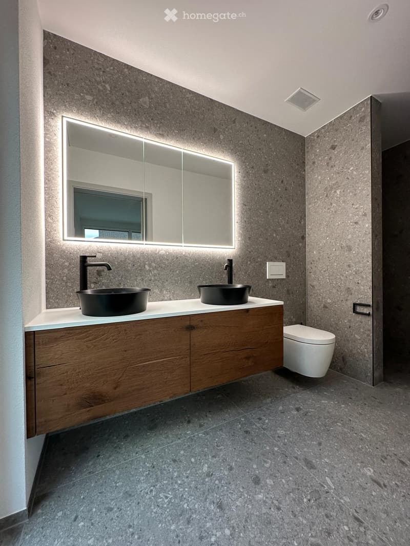 Modern bathroom with dark stone walls, two black sinks, wooden cabinet, large mirror with LED lighting, and a toilet.