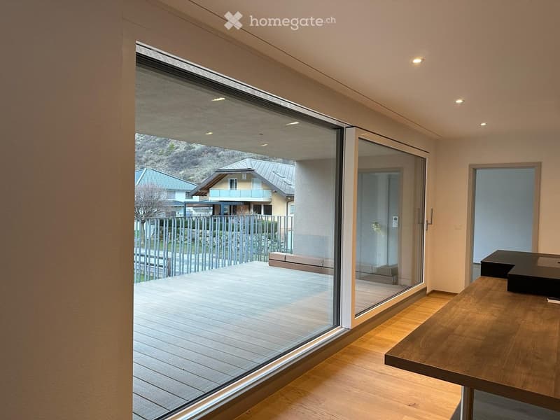 Modern interior with large glass sliding doors leading to a wooden deck, a wooden table, and a view of the outdoor area and neighboring houses.