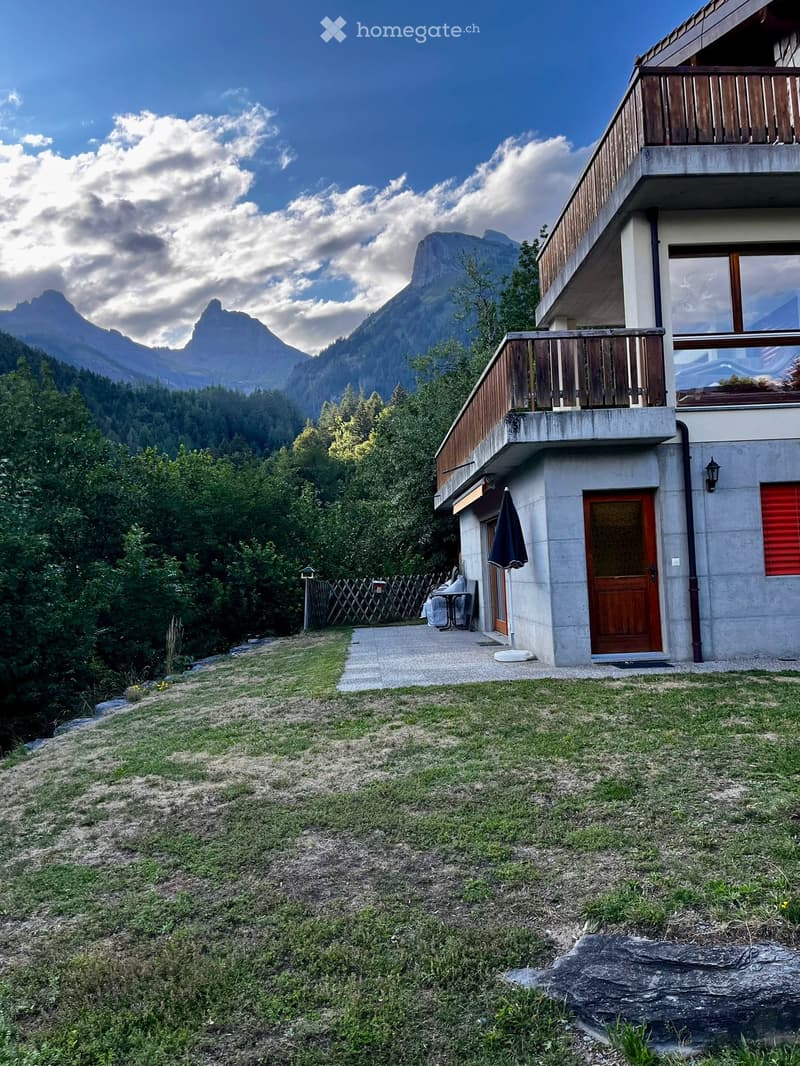 Two-story house with a balcony, wooden door, and red shutters, surrounded by a grassy yard and trees, with a mountain view in the background.