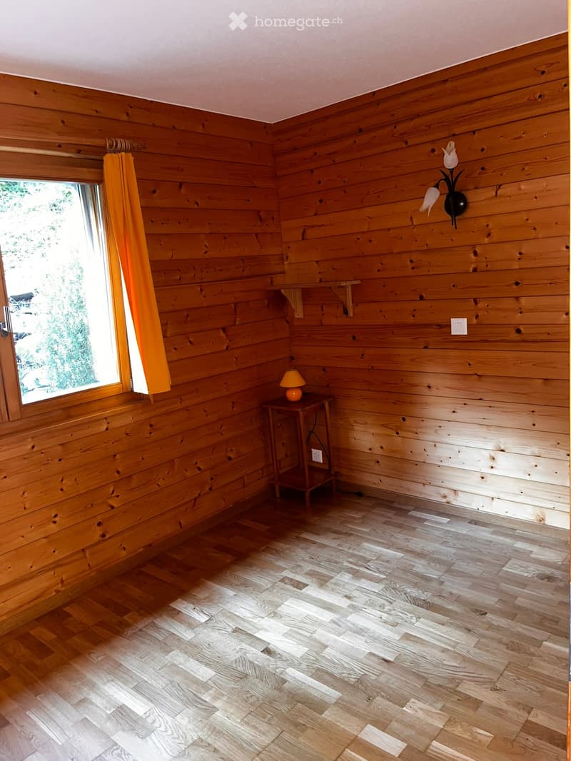Empty room with wooden walls and flooring, a small table with a lamp, and a window with orange curtains.