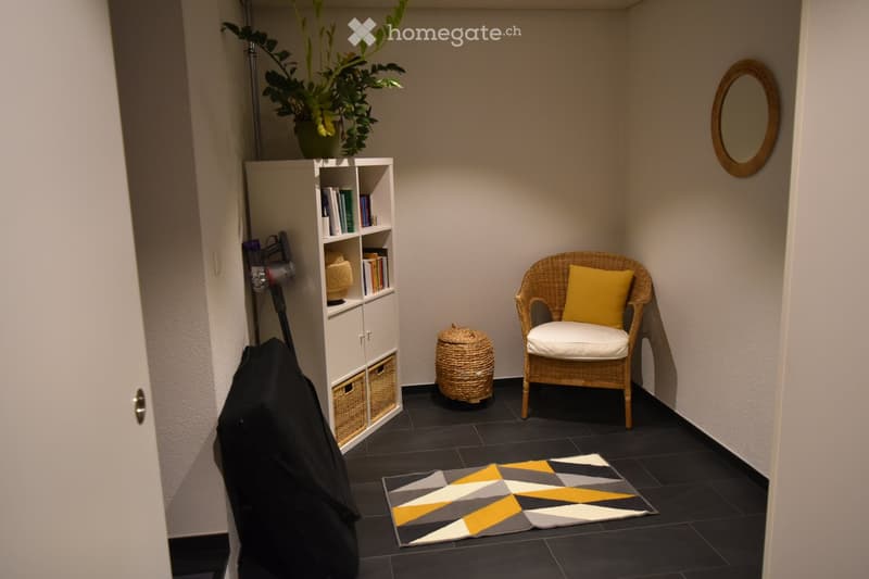 A small, cozy room with a wicker chair, a shelf with books and decorative items, a round mirror on the wall, a potted plant, and a patterned rug on the floor.