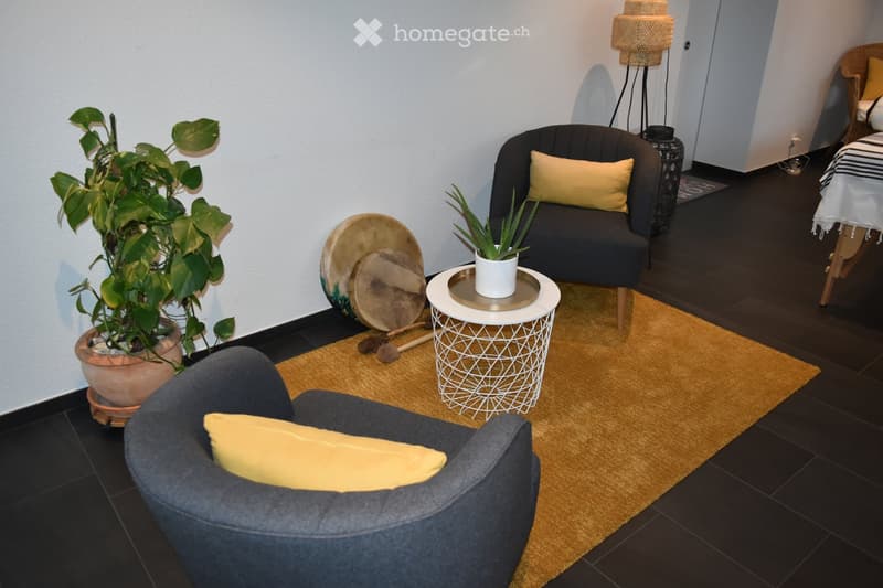 A cozy living room with dark wooden flooring, two armchairs with yellow cushions, a small white table with a plant, and a large potted plant. A drum set is placed near the wall.