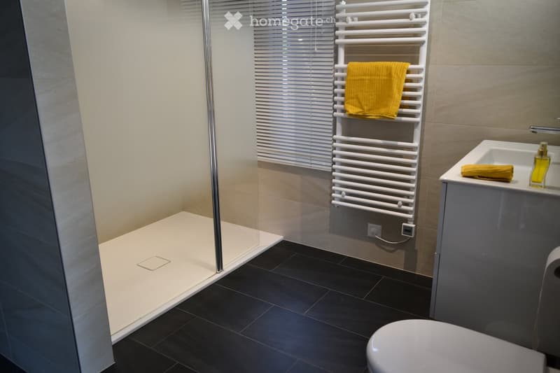 Modern bathroom with a white tiled shower area, black tiled floor, white radiator with a yellow towel, a white sink with a faucet, and a toilet.