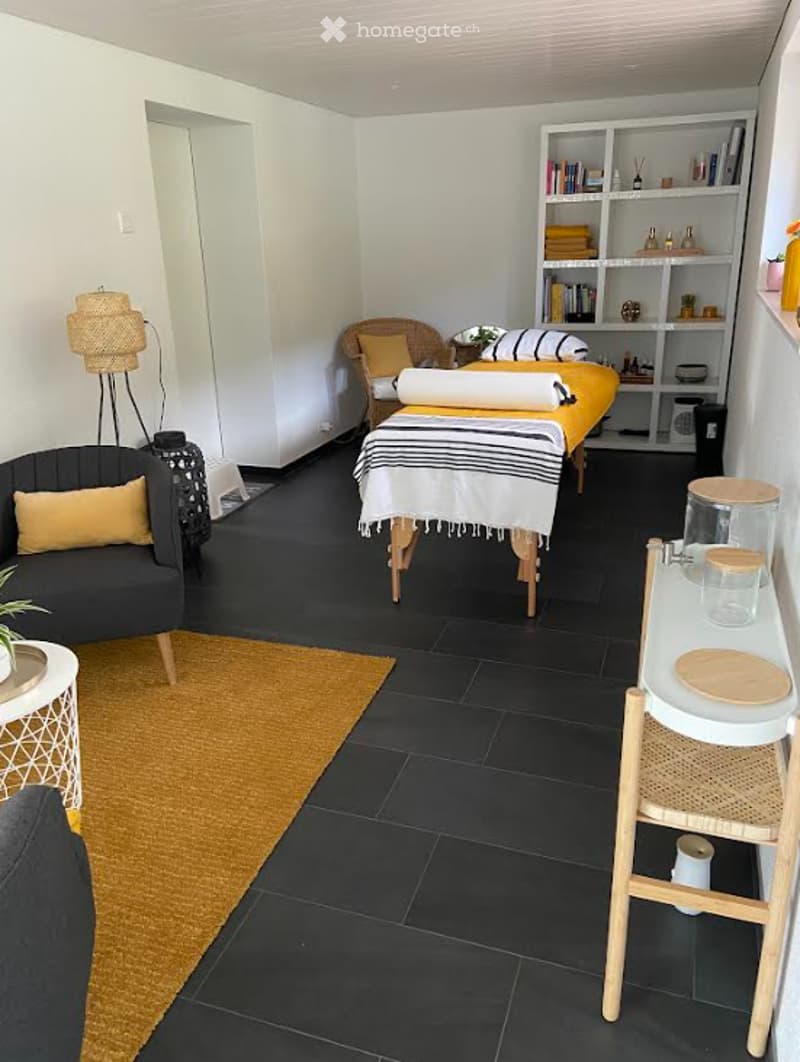 A modern, well-lit room with a black tiled floor, a grey sofa with yellow cushions, a small round table with a plant, a wicker chair with a yellow cushion, a massage table with a striped blanket, and a white shelving unit with books and decorative items.