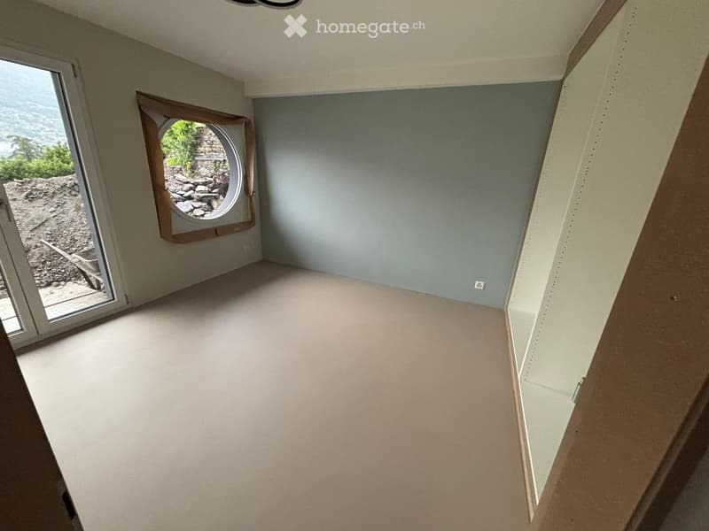 Empty room with light blue walls, white flooring, and a round window with a wooden frame. The room has a slanted ceiling and a door leading outside.