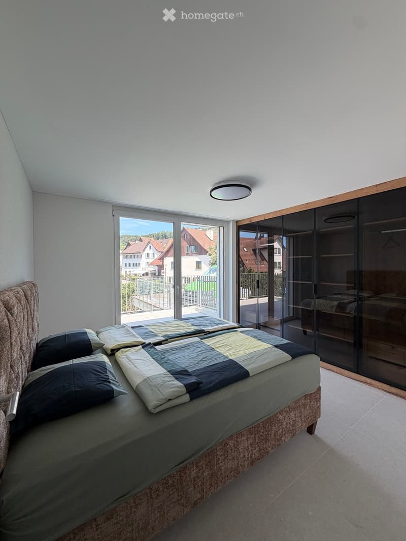 Modern bedroom with a large bed, patterned bedding, and a sliding wardrobe. Large windows offer a view of the neighborhood.