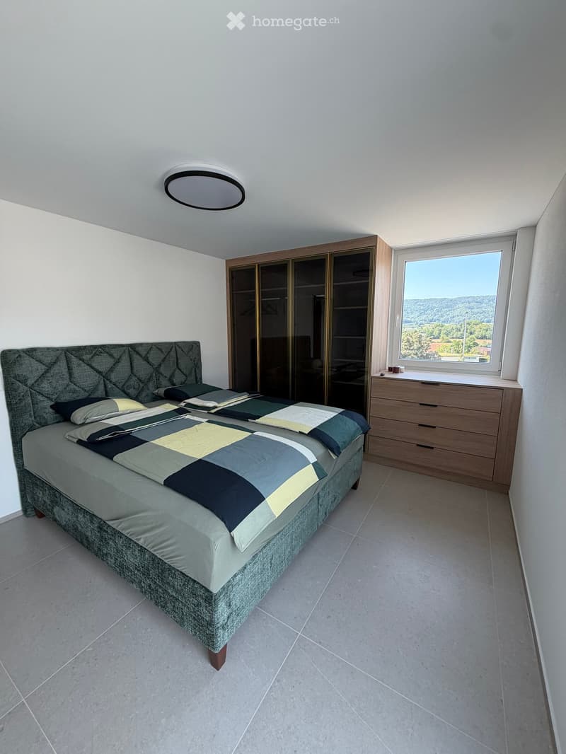 Modern bedroom with a large bed, patterned bedding, a wardrobe, and a nightstand near a window with a scenic view.