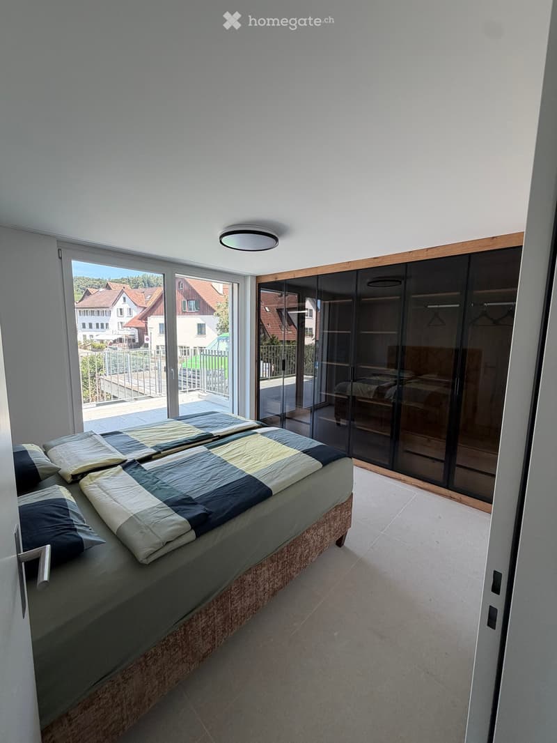 Modern bedroom with a large bed, pillows, and a wardrobe with glass doors. Large windows offer a view of the neighborhood.