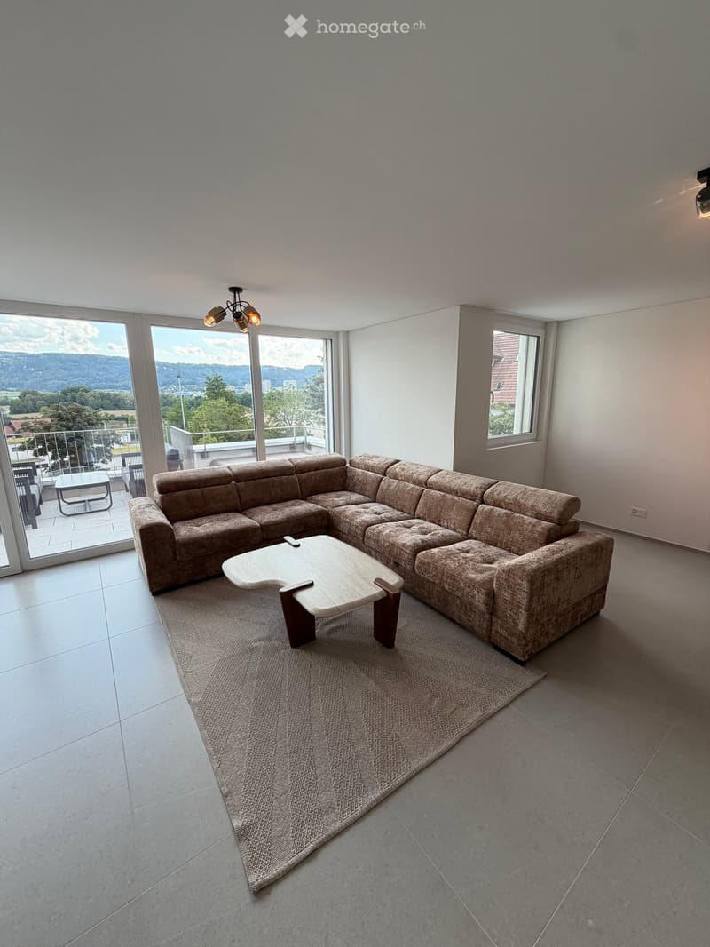 Modern living room with large windows offering a scenic view, featuring a sectional sofa, coffee table, and a small table with chairs on a balcony.