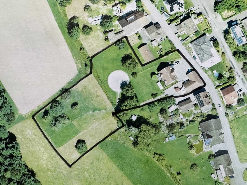Aerial view of a residential area with multiple houses, gardens, and a large open field. The property includes a fenced boundary, trees, and a circular driveway.