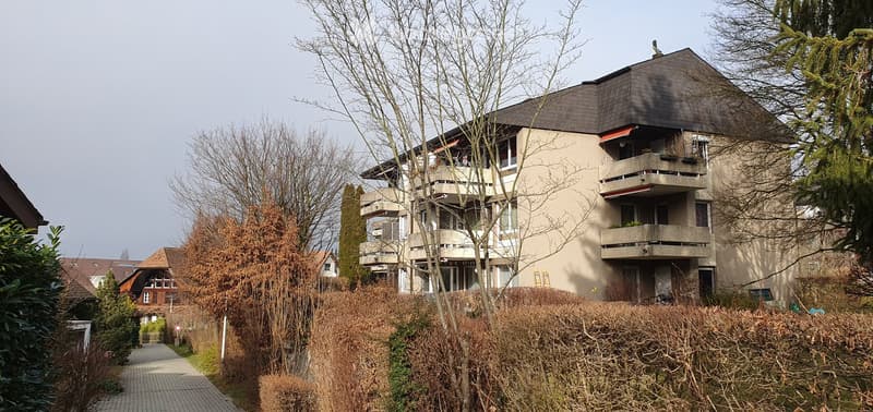 Modern multi-story apartment building with balconies, surrounded by trees and shrubs.