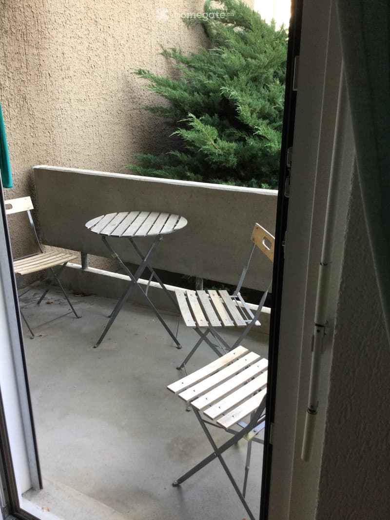 Balcony with a small round table and three wooden folding chairs, overlooking a green bush.