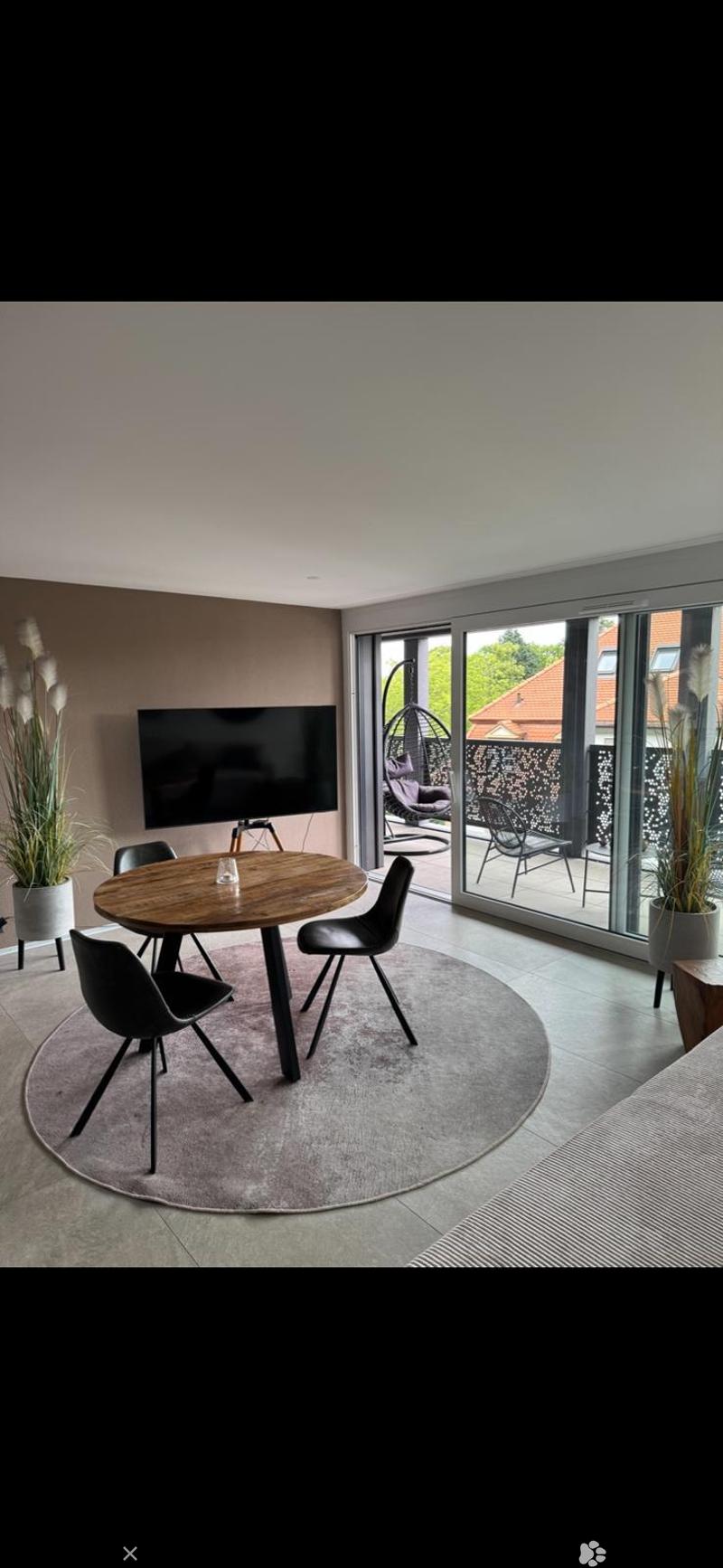 Modern dining area with a round wooden table, black chairs, flat-screen TV, and large glass sliding doors leading to a patio with outdoor seating and plants.