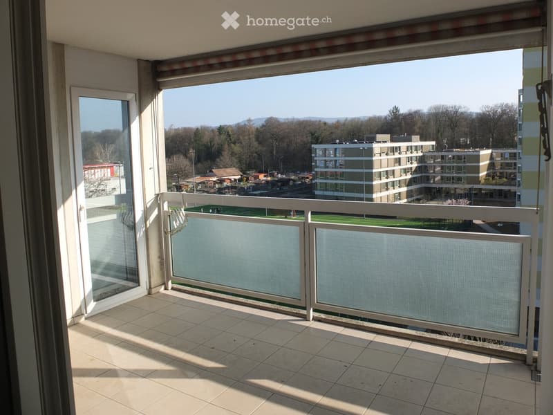 Balcony with tiled floor, white railing, and a view of a green field, trees, and buildings.