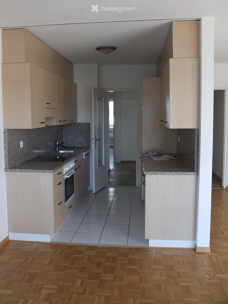 Modern kitchen with light wooden cabinets, granite countertops, and a tiled floor. The kitchen features a sink, oven, and built-in cabinets. The adjacent area shows a hallway with wooden flooring.
