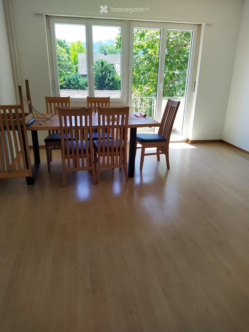 Dining room with wooden floor, dining table, and chairs, large windows with a view of greenery.