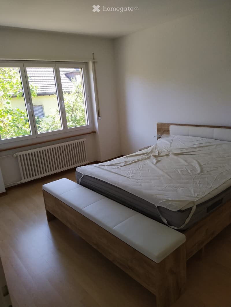 Bedroom with a wooden bed, a bench at the foot of the bed, and a large window with a view of greenery.