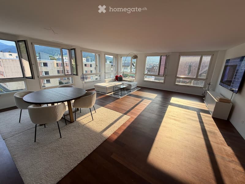 Bright, open living space with large windows offering a scenic view, featuring a round dining table with chairs, a white sofa with an ottoman, a glass coffee table, and a television on a stand.