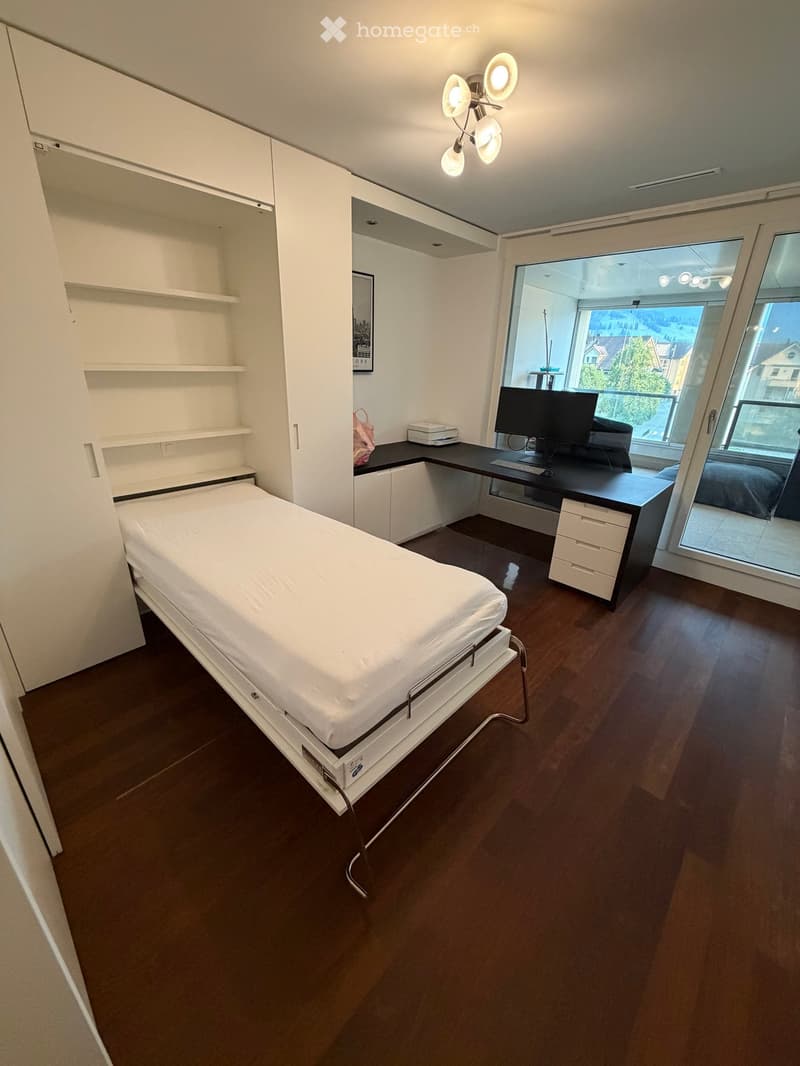 Modern bedroom with a single bed, desk, chair, computer, and shelves. Large windows offer a view of the city.