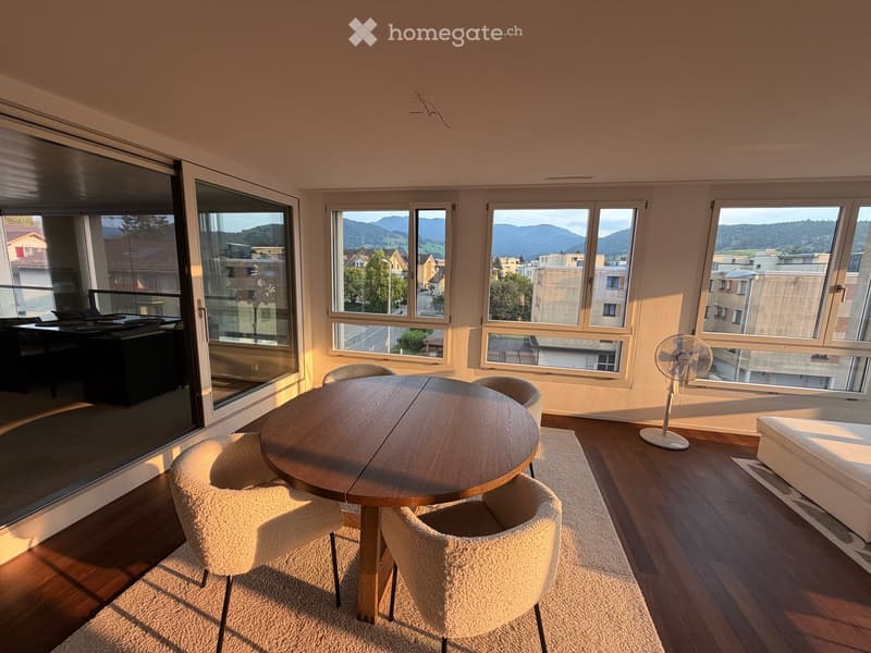 Bright room with large windows and a balcony, featuring a round wooden table with four chairs, a fan, and a view of the city and mountains.