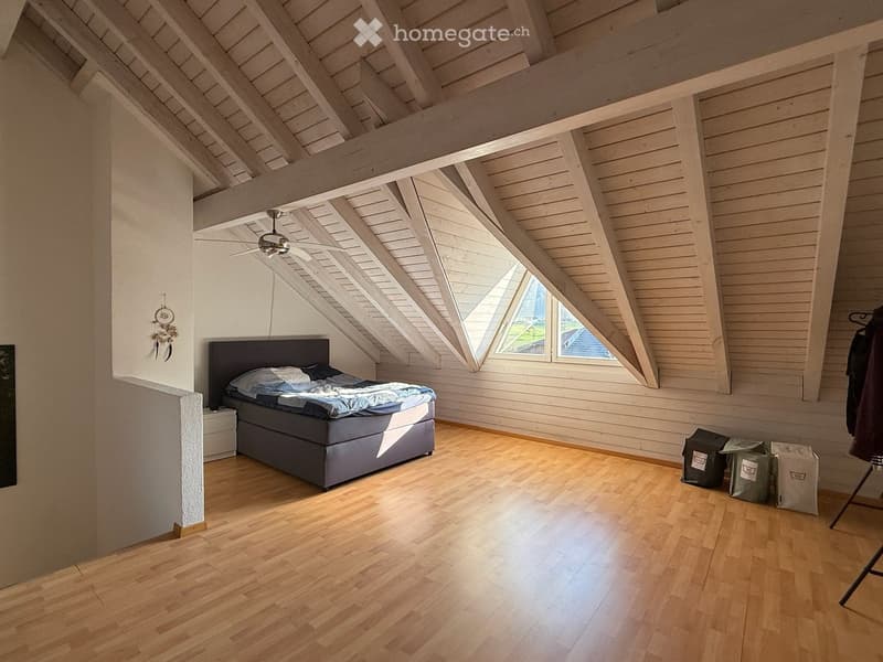 Bright, airy attic bedroom with wooden floors, exposed wooden beams, and large skylight windows. Features a double bed with blue bedding, a nightstand, and a ceiling fan.