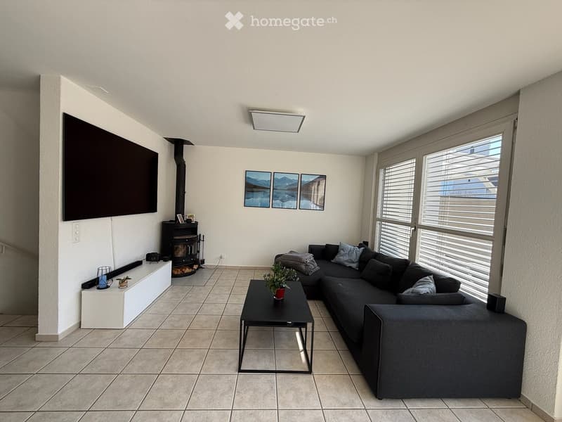 Modern living room with a dark grey sectional sofa, black coffee table, fireplace, large TV, and large windows with blinds.