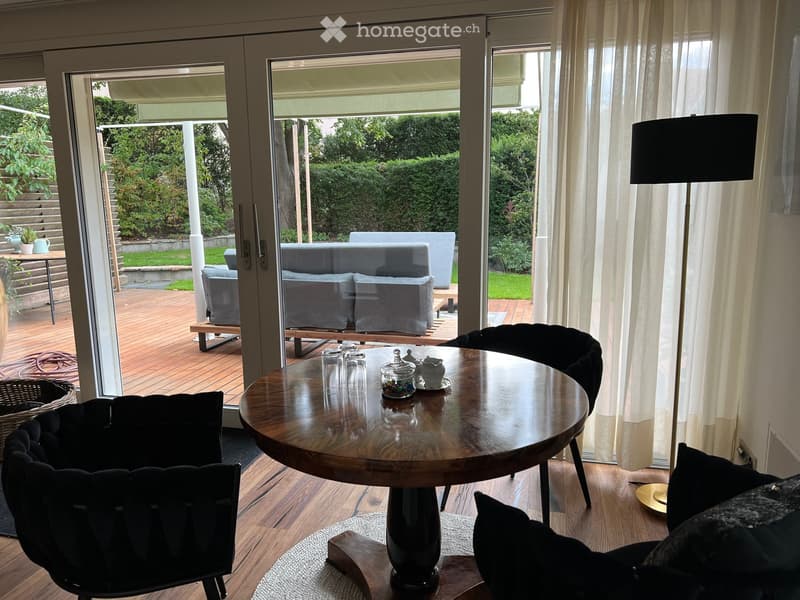 Bright living room with large windows and sliding doors leading to a wooden deck. Features a round wooden coffee table, black leather chairs, a grey sofa, and a floor lamp.