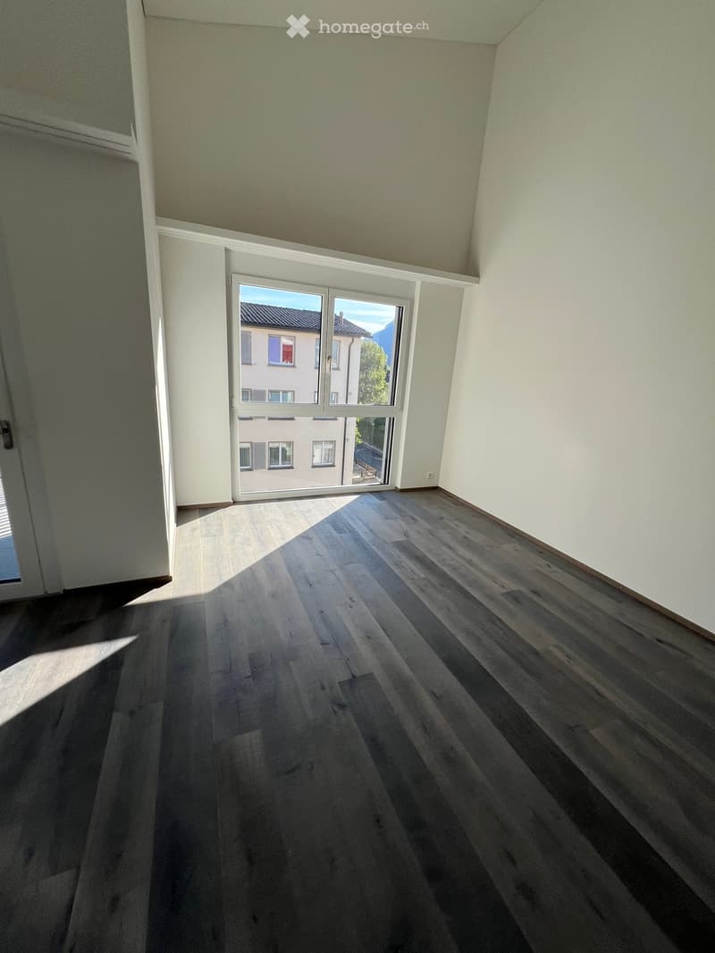 Empty room with wooden flooring and large windows, allowing natural light to enter. The walls are painted in a light color, and the ceiling has a slightly sloped design.