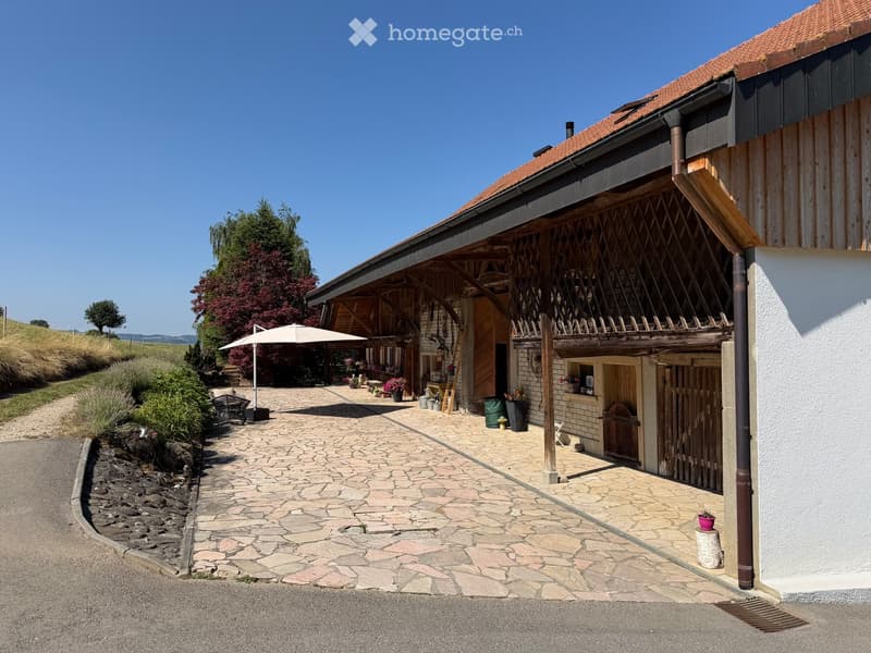 A rustic, two-story wooden building with a red-tiled roof, stone-paved courtyard, and outdoor seating area with an umbrella.