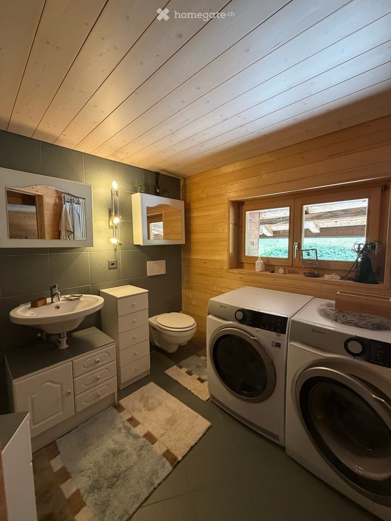 Cozy attic bathroom with wooden walls and ceiling, featuring a sink, toilet, mirror, cabinets, and a washer/dryer unit.