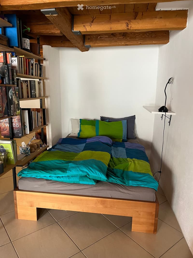 A small bedroom with a wooden bed, colorful bedding, a bookshelf filled with books, and a small table with a lamp.